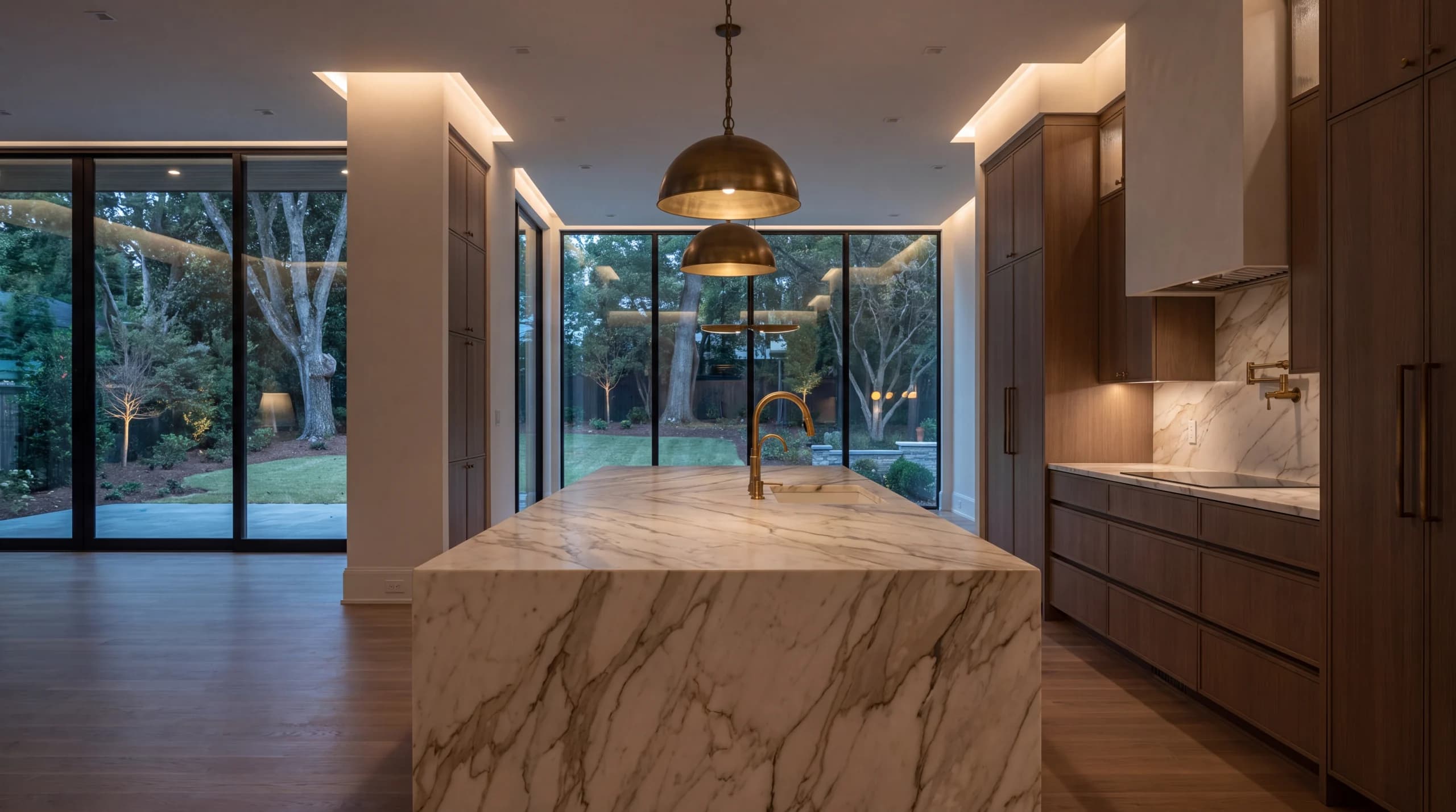 Dusk kitchen with a marble island, walnut cabinets, and brass pendants over the sink.
